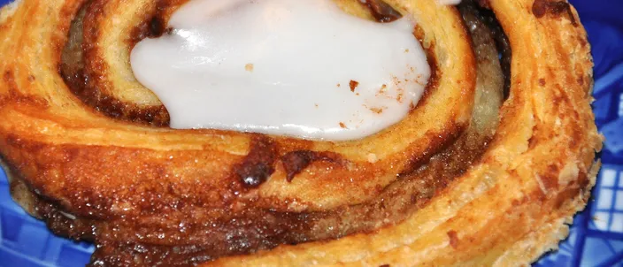 Close-up of a kanelsnegl, a spiral of flaky laminated pastry with a dark cinnamon filling and a pool of white icing in the center, on a blue plate