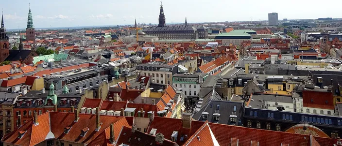 Red-tiled roofs of Copenhagen's old town seen from above, with green copper spires on the left and Christiansborg Palace's dark tower rising in the middle distance