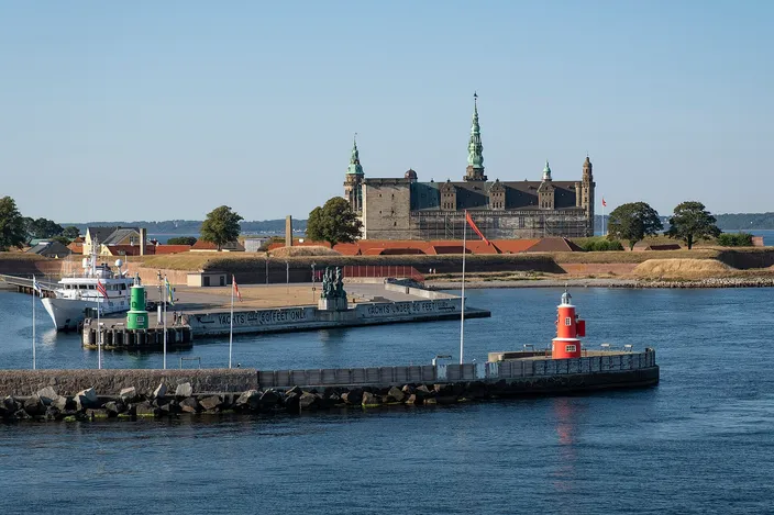 Kronborg Castle's sandstone walls and green spires across Helsingør's harbor, with moored boats and a red harbor light in the foreground