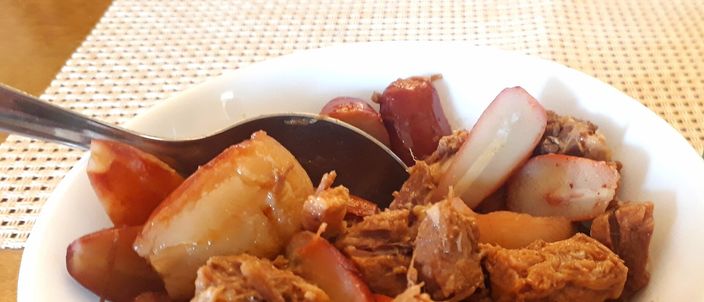 A white bowl of entrevero, holding pale boiled pinhao seeds among pieces of braised meat and onion, with a spoon resting in it on a woven mat.