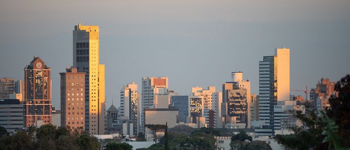Curitiba's mid-rise downtown skyline lit warm gold at sunset, tower blocks rising above a dark band of trees under a soft dusk sky