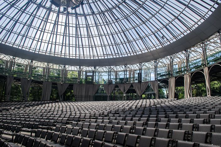 The inside of a theatre roofed by a shallow dome of thin steel tubes and glass, ranks of dark folding seats filling the floor and green trees showing through the transparent walls behind the stage