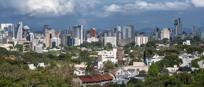 Curitiba's downtown towers strung across the middle distance in bright sun under a bank of dark storm cloud with blue sky behind it, a low suburb of white houses and red tiled roofs below them and a dark green belt of trees across the foreground