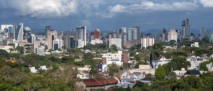 Curitiba's downtown towers strung across the middle distance in bright sun under a bank of dark storm cloud with blue sky behind it, a low suburb of white houses and red tiled roofs below them and a dark green belt of trees across the foreground