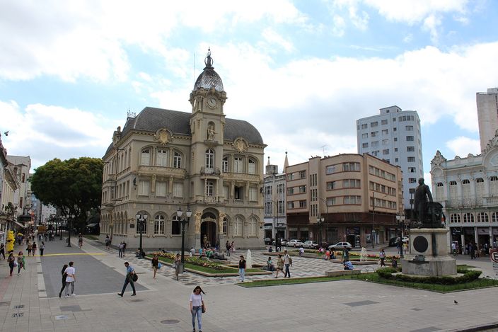 An ornate cream-coloured 1910s civic building with a clock tower and a small dome, standing at the corner of a paved square where people sit on steps and walk past low hedges, taller modern blocks behind