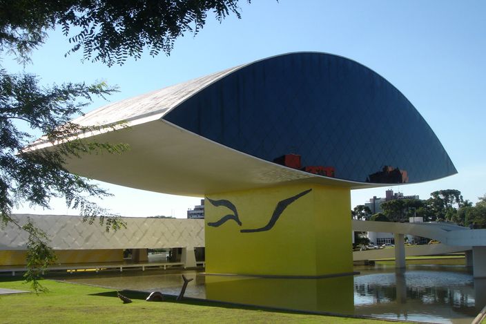 A museum annexe shaped like a huge horizontal eye — a white concrete shell with a dark blue glazed lens set in it — raised on a single yellow pillar above a reflecting pool and a lawn, with a long low white building behind it