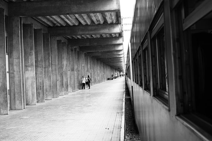 Black-and-white photograph of a passenger train standing alongside an empty platform under a long concrete colonnade that recedes into the distance, two small figures walking towards the camera far down it