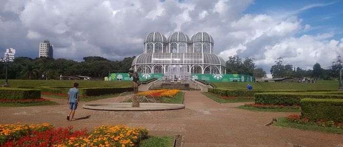 A long formal garden of clipped hedges and beds of orange and yellow flowers running away from the viewer to a white glass-and-iron greenhouse with three curved domes, under a tall bank of white cumulus cloud