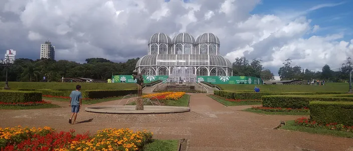 A long formal garden of clipped hedges and beds of orange and yellow flowers running away from the viewer to a white glass-and-iron greenhouse with three curved domes, under a tall bank of white cumulus cloud