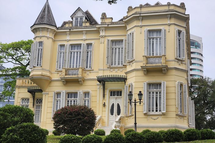 A yellow two-storey belle-époque mansion with a slate-roofed corner turret, grey shutters and manicured hedges, a modern glass tower rising behind