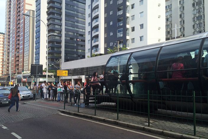 A cylindrical glass tube bus station on a Curitiba busway with commuters queuing to board an articulated bus, apartment towers behind