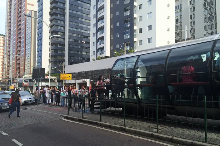A cylindrical glass tube bus station on a Curitiba busway with commuters lining up to board an articulated bus, apartment towers behind