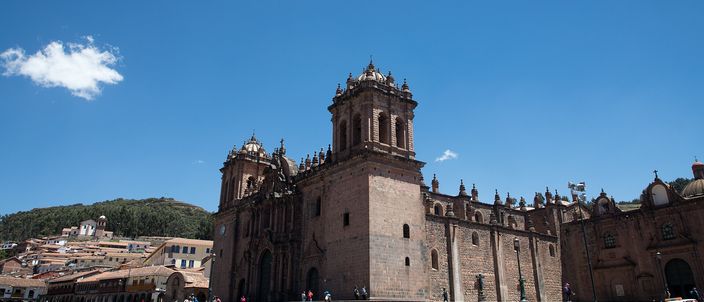 Cusco Cathedral seen from the corner of the Plaza de Armas in strong sun: a broad stone facade between two square bell towers, a long flight of steps below it, the arcaded colonnades of the square running away to the left and a green hill behind