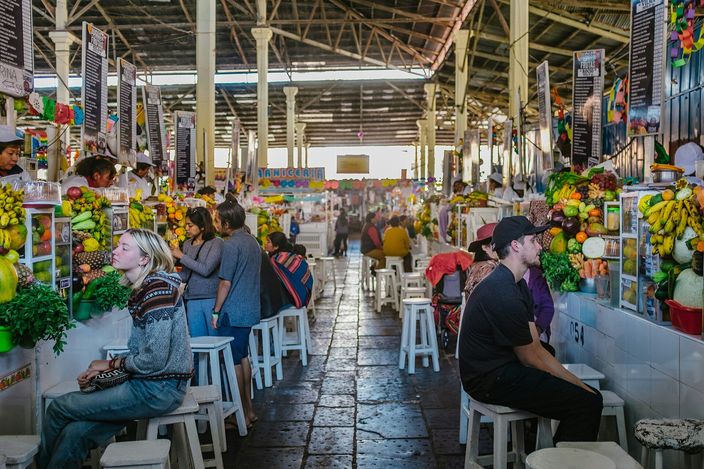 The interior of Cusco's San Pedro market: a wide aisle under a high metal-truss roof running between counters of stacked fruit, hanging bananas and juice stalls, with customers sitting on white plastic stools at the counters on both sides