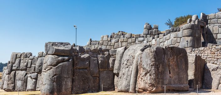 A long run of the megalithic zigzag wall at Sacsayhuamán in strong sun against a clear blue sky: grey limestone blocks, several of them taller than a person, fitted together without mortar in irregular interlocking shapes above a strip of gravel