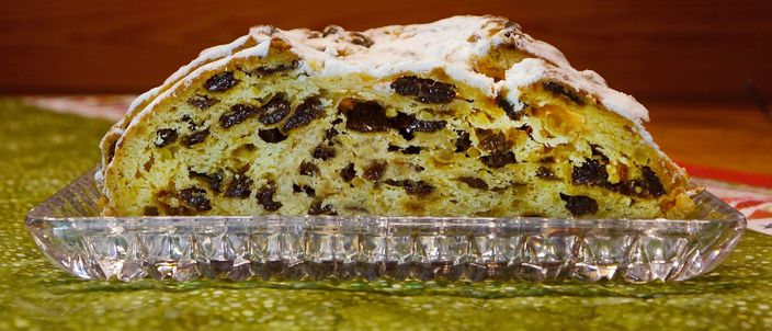 A cut half of Dresdner Christstollen on a glass dish, its dense crumb packed with dark raisins and candied peel under an icing-sugar-dusted crust.