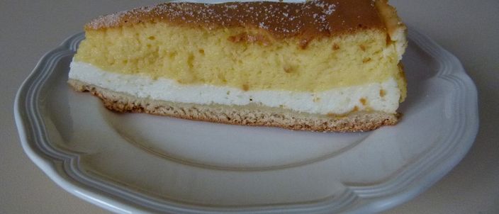 A slice of Dresdner Eierschecke seen from the side on a white plate, showing three distinct layers: a golden set egg custard on top, a white quark layer beneath, and a thin pale base.