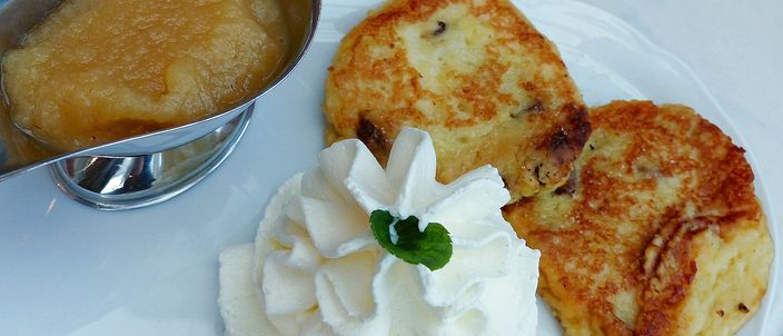 Two golden pan-fried Quarkkeulchen on a white plate, their surfaces flecked with raisins, served with a small jug of apple sauce and a swirl of whipped cream topped with a mint leaf.