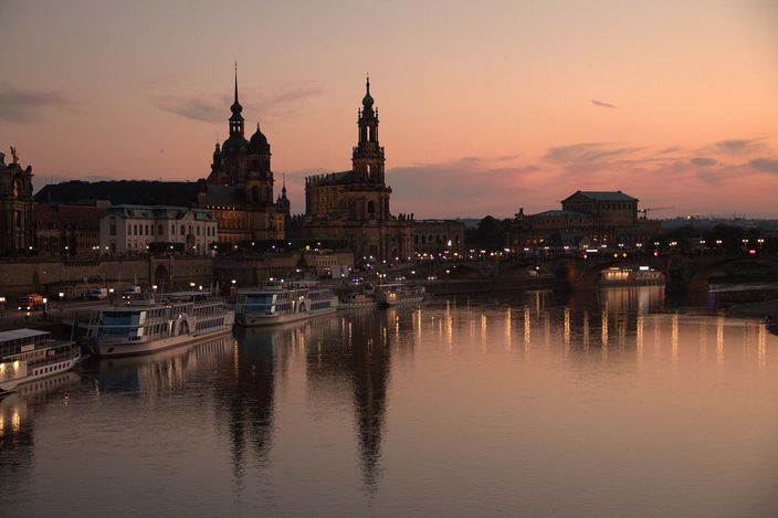 The Dresden Altstadt in silhouette across the Elbe at dusk under a pink and orange sky: the tower of the Katholische Hofkirche and the spire above the castle rising behind the long frontage of the Brühlsche Terrasse, a row of lit street lamps casting broken reflections on the water, white paddle-steamers moored along the far quay and the stone arches of the Augustusbrücke on the right.