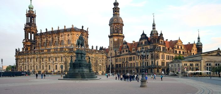 Theaterplatz in Dresden seen at ground level across the empty cobbles: the Katholische Hofkirche and its openwork bell tower on the left, the Residenzschloss with the clock-faced Hausmannsturm and red roofs on the right, the bronze equestrian statue of King Johann in the middle, small groups of people walking under a pale morning sky.