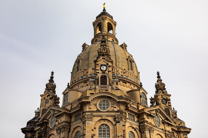 The dome of the Dresden Frauenkirche seen from below against a pale grey sky: pale honey sandstone blotched with soot-black salvaged blocks, a clock set into the drum, four curved gables with scrolled volutes around it, and the stone lantern on top carrying a gilded cross.