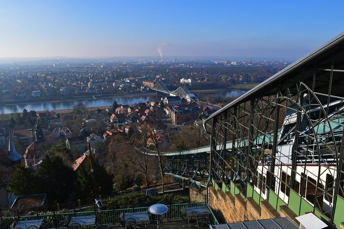 The view from the roof of the Loschwitz upper station: the green steel truss and glazed upper terminal of the Dresden suspension railway fill the right of the frame, and beyond it the wooded slope drops to villa roofs, the Elbe curving away west with the pale blue steel arch of the Blaues Wunder bridge across it, and the flat hazy city stretching to the horizon under a winter sun.