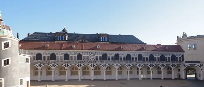 The Stallhof in Dresden: a long two-story Renaissance range closing an empty sunlit courtyard, with a white ground-floor arcade of round arches on squat columns, a sgraffito-patterned upper facade of black and cream panels, a dark slate roof with dormers and a small lantern, and a deep blue cloudless sky above.