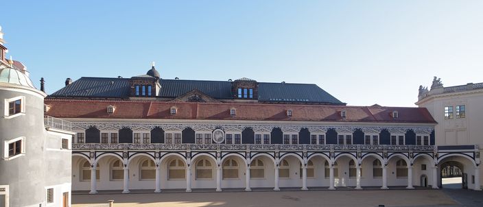 The Stallhof in Dresden: a long two-storey Renaissance range closing an empty sunlit courtyard, with a white ground-floor arcade of round arches on squat columns, a sgraffito-patterned upper facade of black and cream panels, a dark slate roof with dormers and a small lantern, and a deep blue cloudless sky above.
