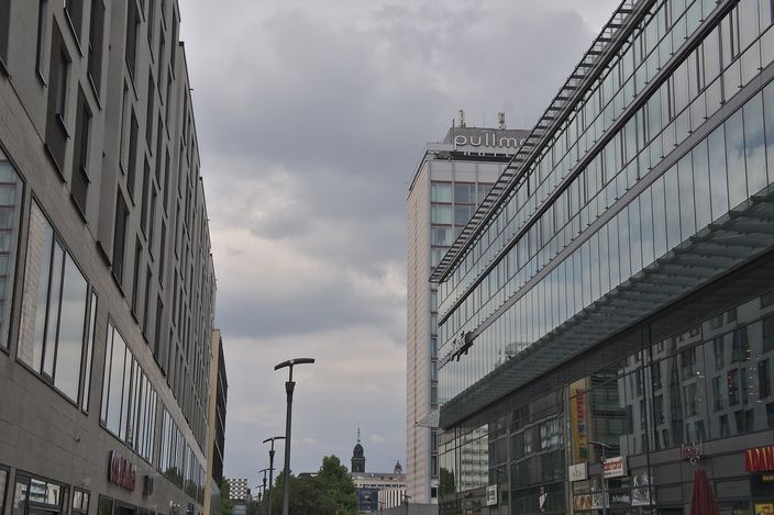 A wide modern shopping street in Dresden lined with glass-fronted buildings, the tall Pullman hotel tower rising at the end under a grey sky, with a baroque church spire glimpsed low between the blocks.