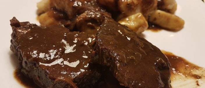 Slices of dark, fork-tender braised beef napped with a glossy deep-brown sauce, served with small potato gnocchi on a white plate.