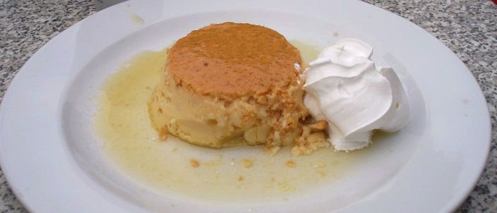 A rožata: a caramel-topped baked custard, unmolded on a plate in a pool of caramel with a spoon of cream.
