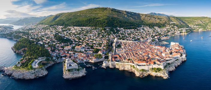 Aerial panorama of Dubrovnik's walled Old Town on its rocky peninsula, terracotta roofs ringed by pale stone walls jutting into the deep-blue Adriatic, with the wooded coast and Mount Srđ rising behind in warm evening light.