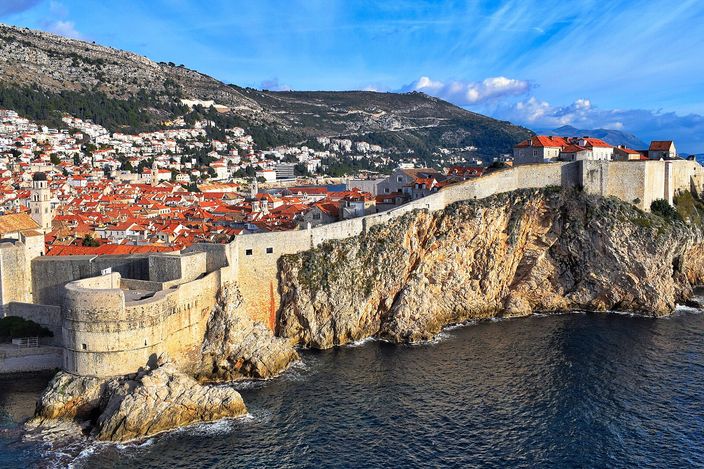 Dubrovnik's city walls running along the top of a sea cliff, pale stone ramparts and a round tower above the Adriatic, with the Old Town's terracotta roofs and a green hillside rising behind.