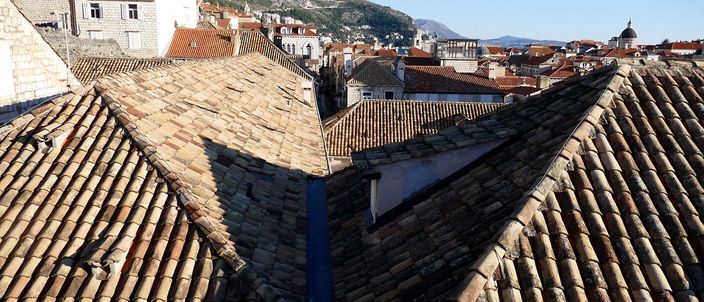 Terracotta pantile roofs of Dubrovnik's Old Town seen from the walkway on top of the city walls, ridge lines converging under a clear blue sky with a wooded hillside and a church dome behind.