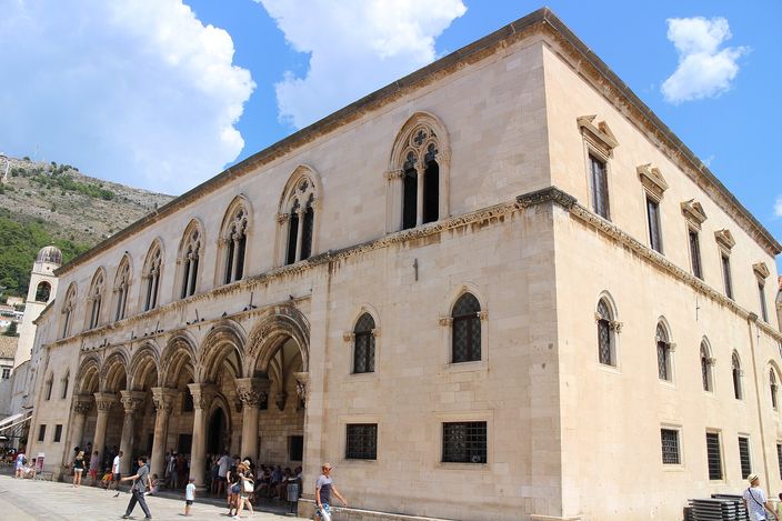 The Rector's Palace in Dubrovnik in full sun: a pale stone block with a ground-floor loggia of carved arches on the left, Gothic windows above, and the cathedral end of the street and a green hillside behind.
