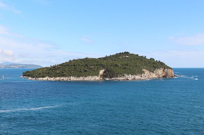 The wooded island of Lokrum seen whole across deep blue water from the Dubrovnik shore, a low dark-green dome of forest with pale rock cliffs at the waterline and open sea and hills behind.