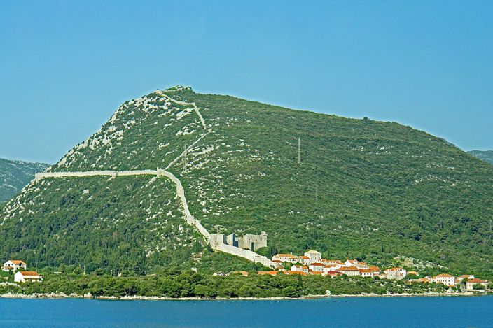 The walls of Ston climbing a steep green hill in white zigzag lines from a small red-roofed village on the shore to the ridge, seen across a strip of calm water.