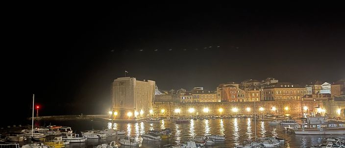 Dubrovnik's old harbour at night: dozens of small moored boats in the foreground, the lit stone quay and the round bulk of St John's Fortress beyond, with the old town's windows glowing above the waterfront.