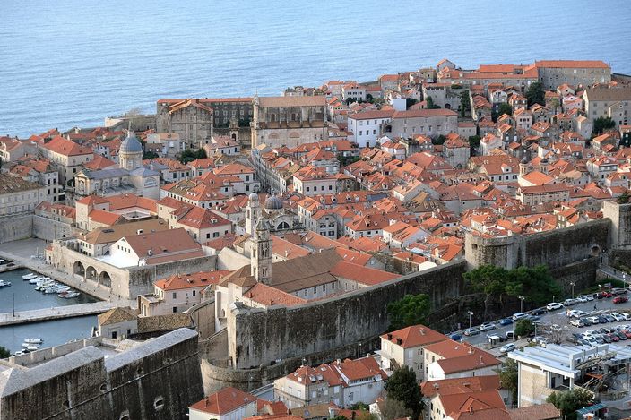 Aerial view over Dubrovnik's Old Town: densely packed orange terracotta roofs, baroque church domes and bell towers, ringed by pale stone city walls with the harbour and Adriatic on the left.