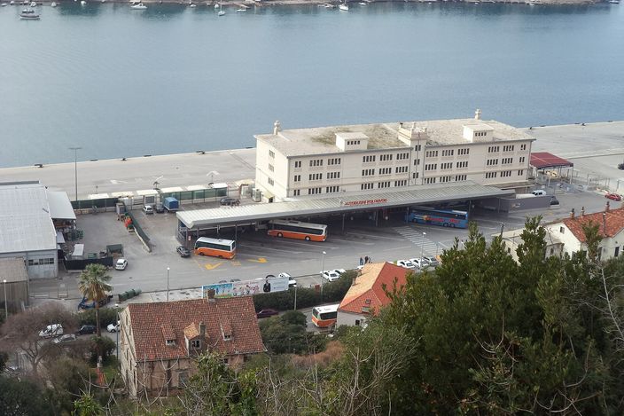Dubrovnik's Gruž bus station from above: a low cream building with orange-and-white city buses parked at bays, cars in the forecourt, the harbour water and wooded far shore behind.