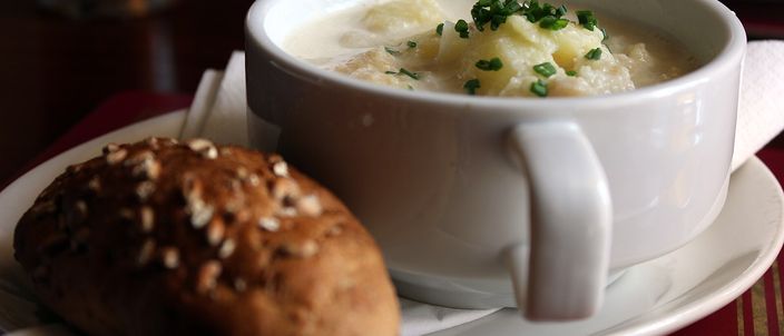A white bowl of pale, flecked Cullen skink showing chunks of fish and potato and a scatter of chives, served on a plate with a seeded bread roll