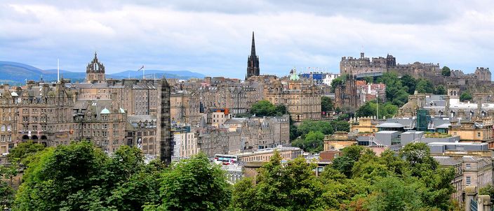 Edinburgh's Old Town skyline seen from Calton Hill: tightly packed stone tenements and spires along the ridge, the crowned tower of the Hub, the Balmoral clock tower at left, and Edinburgh Castle on its rock at right, under a soft grey sky with green trees in the foreground.