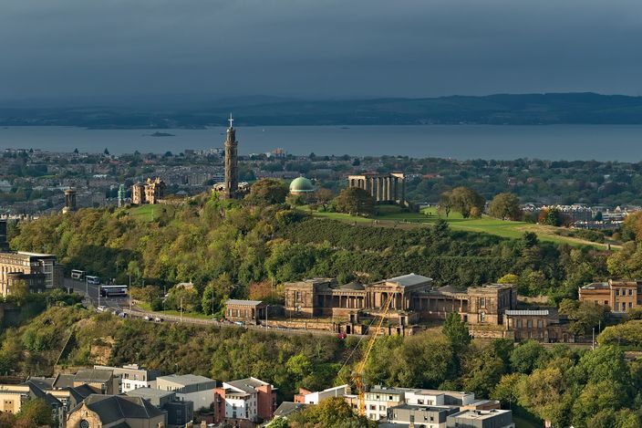 A green hill above a city, carrying a tall stone column topped by a mast, a row of unfinished classical columns, and a domed observatory among trees, with a broad estuary and a far shoreline behind it in low golden light.