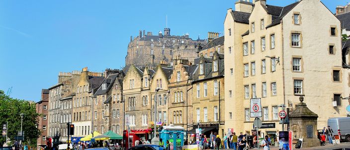 A sunlit open square in Edinburgh lined with tall stone tenements and ground-floor shopfronts, with market stalls, parked cars and people crossing the foreground, and Edinburgh Castle standing on its dark volcanic rock above the rooftops behind.