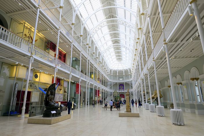 A long, tall museum hall lit by an arched glass roof carried on slender white cast-iron columns, with tiered balconies down both sides, display cases and sculpture standing on the pale stone floor, and a handful of visitors walking through.