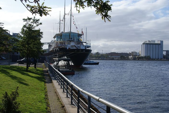A large dark-hulled former royal yacht with a pale superstructure and tall masts, berthed alongside a quay, seen past a grass verge, trees and a metal handrail, with open water and dockside buildings behind.