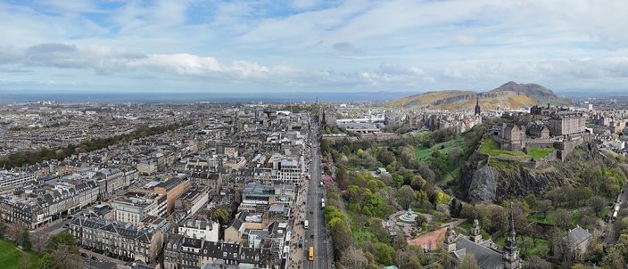 An aerial view over Edinburgh: the grey rooftops and straight streets of the New Town in the foreground, a long avenue running east between them, the wooded castle rock with the castle on top at right, and the green hump of Arthur's Seat on the horizon beyond, under a cloudy sky.
