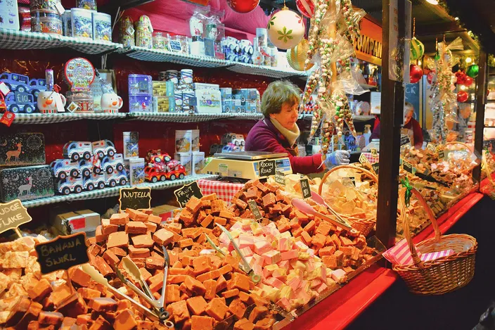 A stallholder behind a counter piled with slabs of fudge in many flavors, labeled by hand, with Christmas tins and ornaments on the shelves above