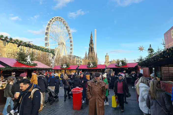 Crowds walking between red-roofed market huts in East Princes Street Gardens on a clear December day, the big wheel and the spire of the Scott Monument behind them