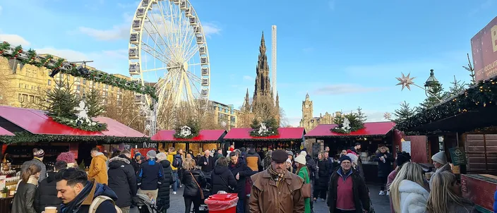 Crowds walking between red-roofed market huts in East Princes Street Gardens on a clear December day, the big wheel and the spire of the Scott Monument behind them